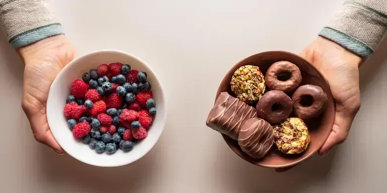 Zwei Hände halten verschiedene Lebensmittel – eine mit einem Schokoriegel und Donuts, die andere mit einer Schale voller Beeren und Nüsse.