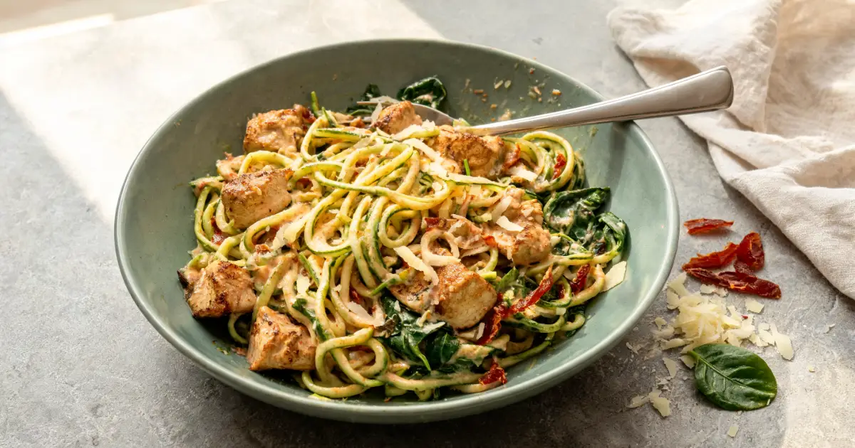 Cremige Zucchini-Nudeln mit gebratenem Hähnchen, Spinat, sonnengetrockneten Tomaten und Parmesan in einer Schale angerichtet.