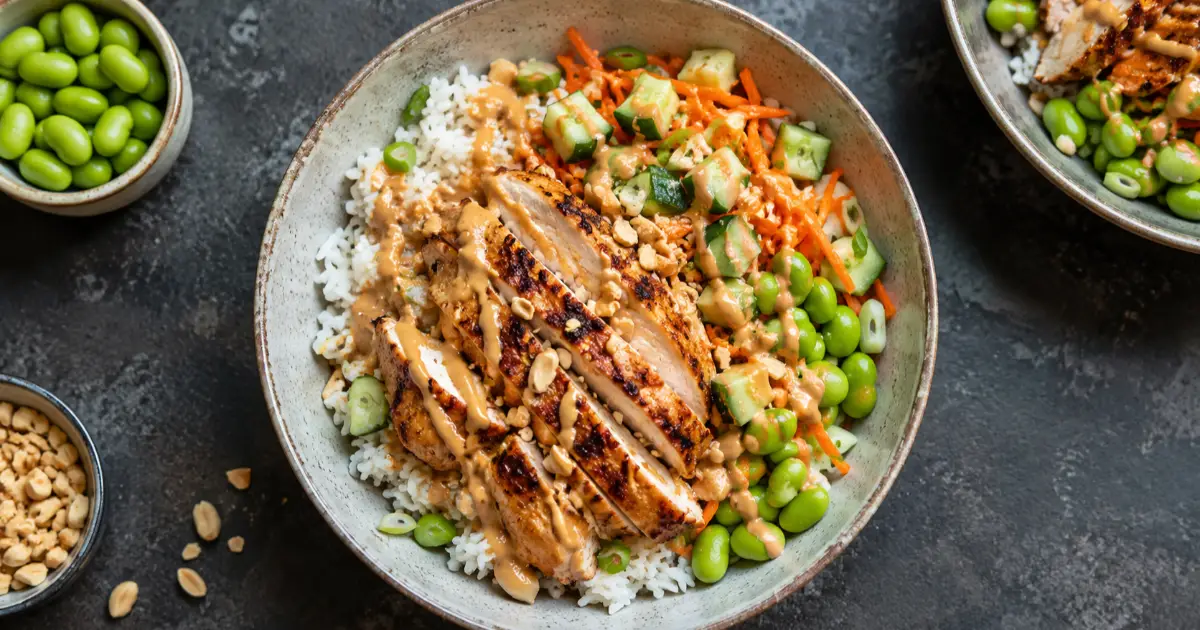 High-Protein Erdnuss-Hähnchen-Bowl mit Reis, Edamame, Gurke, Karotte und cremiger Erdnusssauce.