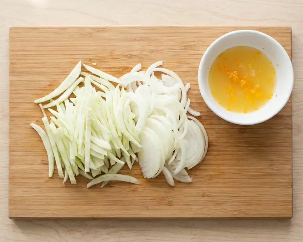 Geschnittener Fenchel und Zwiebel auf Holzbrett mit kleiner Schüssel Orangensaft und Abrieb von oben