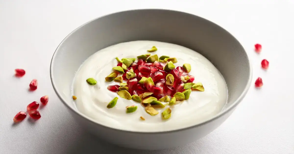 Kokosjoghurt mit Granatapfelkernen und gehackten Pistazien, serviert in einer Schale auf einem weißen Untergrund.