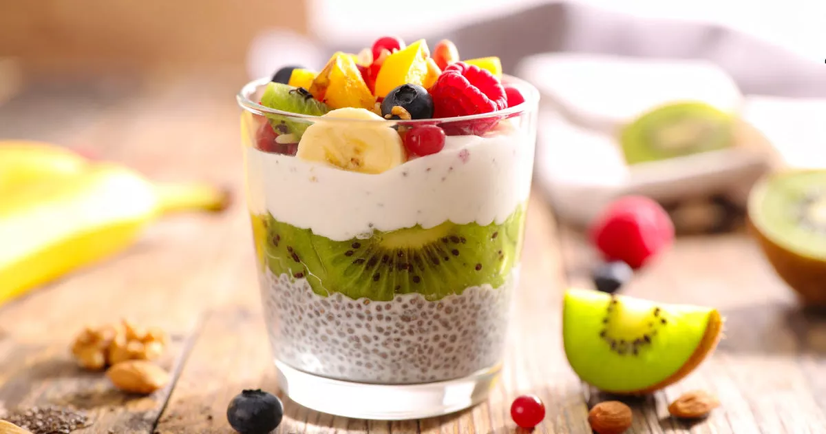 Ein Glas mit Chia-Pudding-Schichten, Proteincreme und frischem Obst, garniert mit Nüssen und Kokosraspeln, auf einem rustikalen Holztisch.